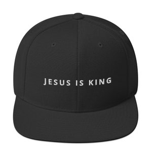 Puede incluir: Gorra de béisbol negra con visera plana. La frase "JESUS IS KING" está bordada en blanco en la parte delantera. La gorra tiene un ajuste estructurado y una visera curva.