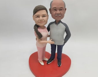May include: A custom-made figurine of a couple standing together on a red heart-shaped base. The woman is wearing a pink outfit and the man is wearing a grey shirt and blue trousers.
