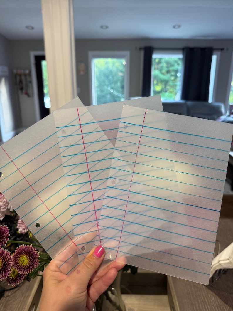 Puede incluir: Un mont&oacute;n de hojas de papel transl&uacute;cido con l&iacute;neas azules y rojas. El papel tiene tres agujeros perforados para usar en una carpeta. Las hojas se sostienen frente a un fondo borroso, con un ramo de flores en la esquina inferior izquierda.