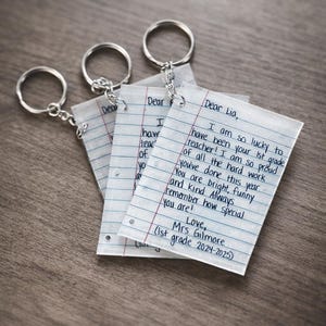 May include: Three keychains with paper-like pendants. Each pendant has a handwritten message with a silver keyring attached. The text on the paper reads "Dear Lia, I am so lucky to have been your 1st grade teacher!"