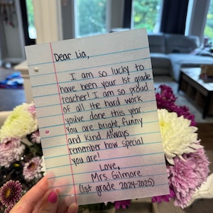 Puede incluir: Una nota manuscrita en papel rayado dirigida a "Lia" por su maestra de 1er grado, la Sra. Gilmore. La nota expresa orgullo y afecto, con texto que incluye "You are bright, funny and kind." Un ramo de flores moradas y blancas se encuentra en el fondo.