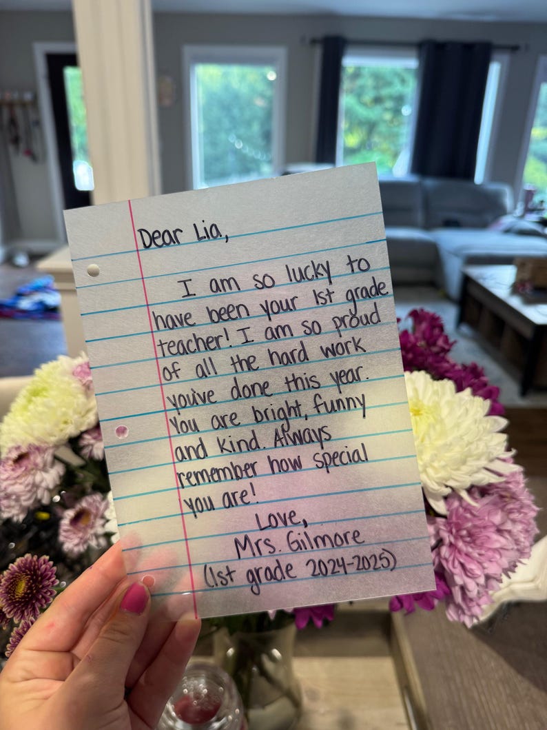Puede incluir: Una nota manuscrita en papel rayado dirigida a "Lia" por su maestra de 1er grado, la Sra. Gilmore. La nota expresa orgullo y afecto, con texto que incluye "You are bright, funny and kind." Un ramo de flores moradas y blancas se encuentra en el fondo.