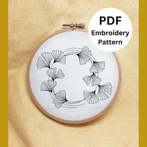May include: A black and white embroidery pattern of a ginkgo leaf wreath on a white fabric in a wooden embroidery hoop. The text "PDF Embroidery Pattern" is on a white circle with a black outline.