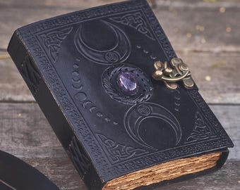 Op de afbeelding: Een zwart lederen dagboek met een reliëf van een drievoudige maan, een centrale paarse edelsteen en een koperen sluiting. Naast het dagboek liggen een zwarte ganzenveer met een zilverkleurig handvat en een klein glazen inktpotje met etiket.