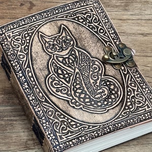 May include: A rectangular leather journal with a dark brown finish featuring an embossed cat design with intricate knotwork patterns. The cover is secured by a metallic bronze-coloured latch and contains white pages visible along the side edge.