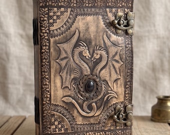 May include: A rectangular brown leather journal featuring an embossed design of two dragons facing each other with a central black stone. The cover includes intricate floral and geometric border patterns, secured by two antique brass metal latches.