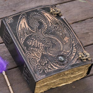 Könnte beinhalten: Ein schwarzes Lederjournal mit einem geprägten Drachen auf dem Einband. Das Journal hat einen Verschluss und gealterte Seiten. Eine lila Federkiel-Feder liegt neben dem Journal auf einer Holzoberfläche.