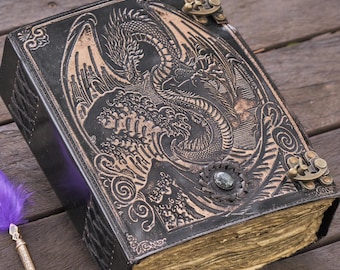 May include: A black leather journal with a dragon embossed on the cover. The journal has a clasp closure and aged pages. A purple feather quill pen rests beside the journal on a wooden surface.