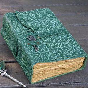 Peut inclure: Un journal relié en cuir vert avec un motif floral décoratif et une fermeture à clé. Un stylo plume en plume de paon est posé sur une surface en bois.