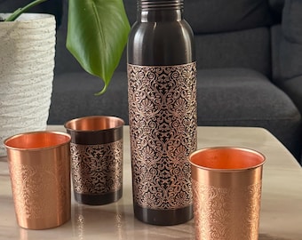 May include: A dark brown metal water bottle with a matching cap and two smaller matching cups. Each piece features an intricate, embossed copper-colored floral pattern that contrasts against the dark finish of the metal exterior.
