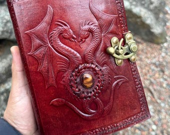 May include: A deep red leather journal featuring an embossed design of two dragons facing each other. The cover includes a central circular braided detail with an embedded brown gemstone and a brass-colored metal latch closure on the right side.