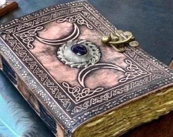 Könnte beinhalten: Ein ledergebundenes Tagebuch mit geprägtem Dreifachmond-Symbol und einem zentralen dunkelvioletten Edelstein. Das Buch hat einen Messingverschluss und gealterte Papierkanten. Eine hellblaue und goldene Federkiel liegt daneben.