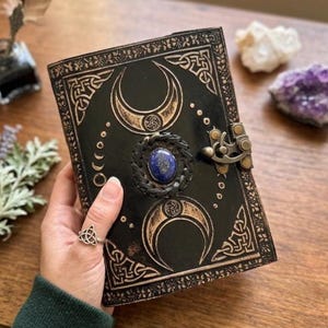 Op de afbeelding: Een in zwart leer gebonden dagboek met in reliëf aangebrachte goudkleurige maanfasen en Keltische knoopranden. De kaft is in het midden voorzien van een gepolijste ovale blauwe lapis lazuli steen in een gevlochten lederen rand, afgesloten met een messing sluiting.