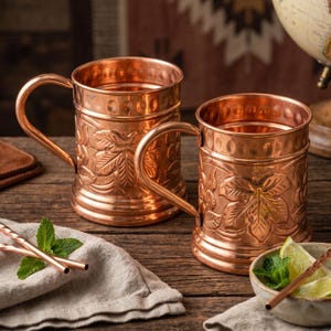 May include: Two polished copper mugs featuring embossed leaf patterns and a decorative band of circular indentations near the rim. Each mug has a curved handle and a cylindrical body, showcasing a warm metallic finish with intricate detailing.