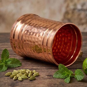 May include: A cylindrical copper mug with a polished finish and a hammered texture on the exterior. The mug features horizontal bands around the top and bottom edges, with a wide opening that reveals the interior surface.