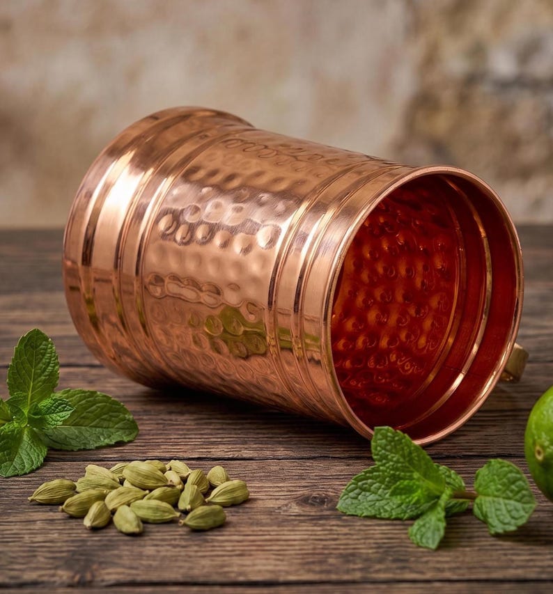 May include: A cylindrical copper mug with a polished finish and a hammered texture on the exterior. The mug features horizontal bands around the top and bottom edges, with a wide opening that reveals the interior surface.