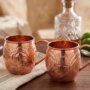 May include: Two copper mugs featuring an intricate embossed floral pattern across the exterior surface. Each mug has a rounded body, a smooth rim, and a curved handle attached to the side. The metal has a polished, reflective finish.