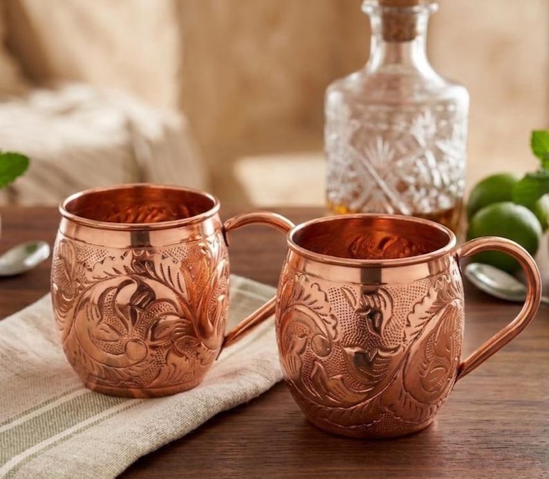 May include: Two copper mugs featuring an intricate embossed floral pattern across the exterior surface. Each mug has a rounded body, a smooth rim, and a curved handle attached to the side. The metal has a polished, reflective finish.