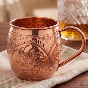 May include: A polished copper mug featuring an intricate embossed floral pattern across its rounded body. The mug has a smooth rim, a sturdy curved handle on the right side, and a metallic finish that reflects light across its textured surface.