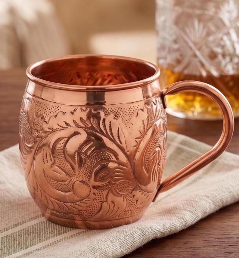 May include: A polished copper mug featuring an intricate embossed floral pattern across its rounded body. The mug has a smooth rim, a sturdy curved handle on the right side, and a metallic finish that reflects light across its textured surface.
