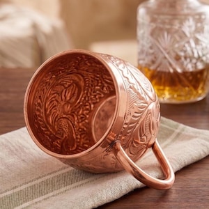 May include: A polished copper mug featuring an intricate embossed floral pattern across its exterior and interior surfaces. The mug has a smooth, rounded handle attached to the side, showcasing a metallic finish and detailed craftsmanship throughout.