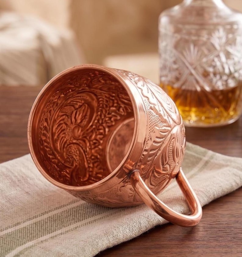 May include: A polished copper mug featuring an intricate embossed floral pattern across its exterior and interior surfaces. The mug has a smooth, rounded handle attached to the side, showcasing a metallic finish and detailed craftsmanship throughout.