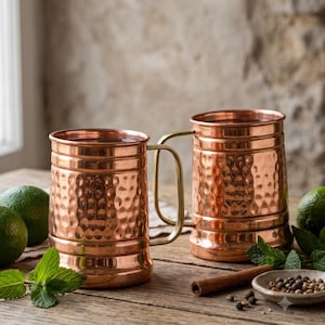 May include: Two copper mugs with a hammered texture and smooth brass handles. The mugs feature a cylindrical body with horizontal bands at the top and bottom. The surface has a polished metallic finish reflecting light across the dimpled exterior.