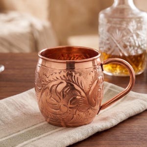 May include: A copper mug with a rounded body and a curved handle, featuring an intricate embossed floral pattern across the exterior surface. The metal has a polished finish that reflects light, highlighting the detailed texture of the design.