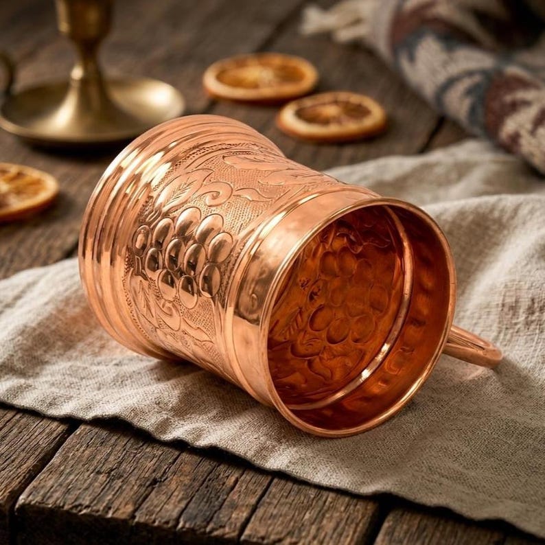 May include: A polished copper mug featuring an embossed design of grape clusters and leaves on its exterior surface. The mug has a cylindrical body with a smooth rim and a handle attached to the side, showcasing a metallic finish and texture.