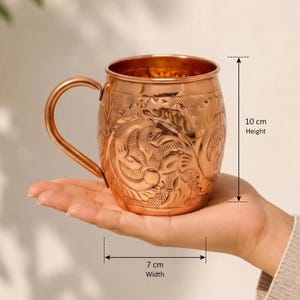 May include: A copper mug featuring an intricate embossed floral pattern and a curved handle. The mug has a height of 10 cm and a width of 7 cm as indicated by the measurement markers. The metallic surface reflects light, highlighting the detailed texture.