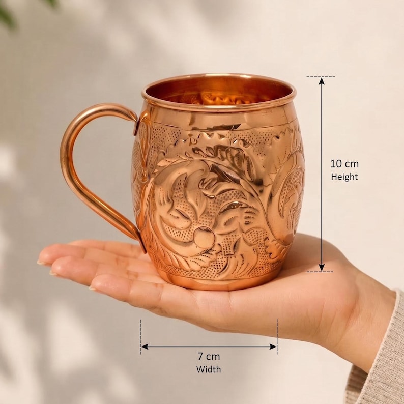 May include: A copper mug featuring an intricate embossed floral pattern and a curved handle. The mug has a height of 10 cm and a width of 7 cm as indicated by the measurement markers. The metallic surface reflects light, highlighting the detailed texture.