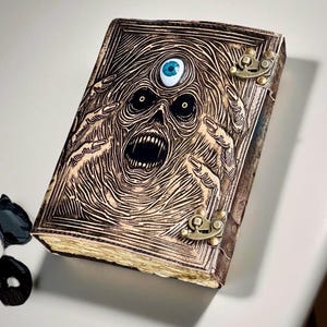 Op de afbeelding: Een bruin, ledergebonden boek met een gebeeldhouwd, schreeuwend gezicht op de omslag. Het gezicht heeft een derde oog en is omgeven door ingewikkelde lijnen. Het boek is vastgezet met messing sluitingen en de pagina's hebben een vintage uitstraling.