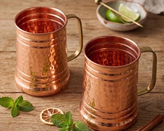 May include: Two hammered copper mugs with vertical cylindrical bodies and attached handles. The mugs feature a textured, dimpled surface finish. Beside the mugs are fresh green mint leaves, a dried citrus slice, and a cinnamon stick.