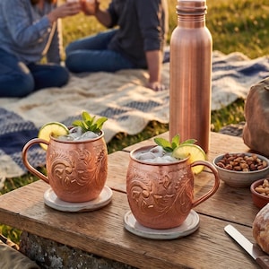 May include: Two copper mugs with intricate embossed floral patterns, each containing ice, a lime slice, and mint leaves. A tall copper water bottle stands nearby on a wooden surface, accompanied by small bowls of nuts and a bread loaf.