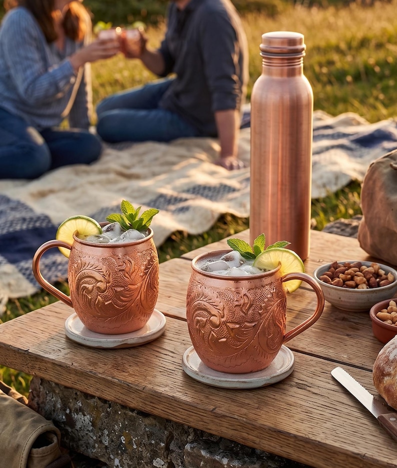 May include: Two copper mugs with intricate embossed floral patterns, each containing ice, a lime slice, and mint leaves. A tall copper water bottle stands nearby on a wooden surface, accompanied by small bowls of nuts and a bread loaf.