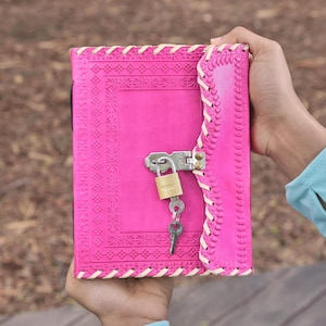 Könnte beinhalten: Ein leuchtend pinkfarbenes Lederjournal mit geprägtem Muster und Schlossverschluss. Das Journal ist mit weißem Faden handgenäht. Ein kleines goldenes Vorhängeschloss und Schlüssel sichern das Journal. Höhe ca. 20 cm.