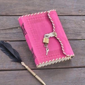 Peut inclure: Un journal en cuir rose vif avec une fermeture à cadenas et clé. Le journal présente une bordure cousue décorative et un motif floral en relief. Un stylo plume à plume noire avec un motif doré est posé à côté.