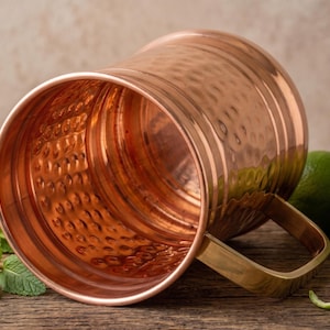 May include: A polished copper mug with a hammered texture on the exterior and interior surfaces. The mug features a smooth, flat-bottomed base and a sturdy, rectangular-shaped handle attached to the side, reflecting a warm metallic sheen.