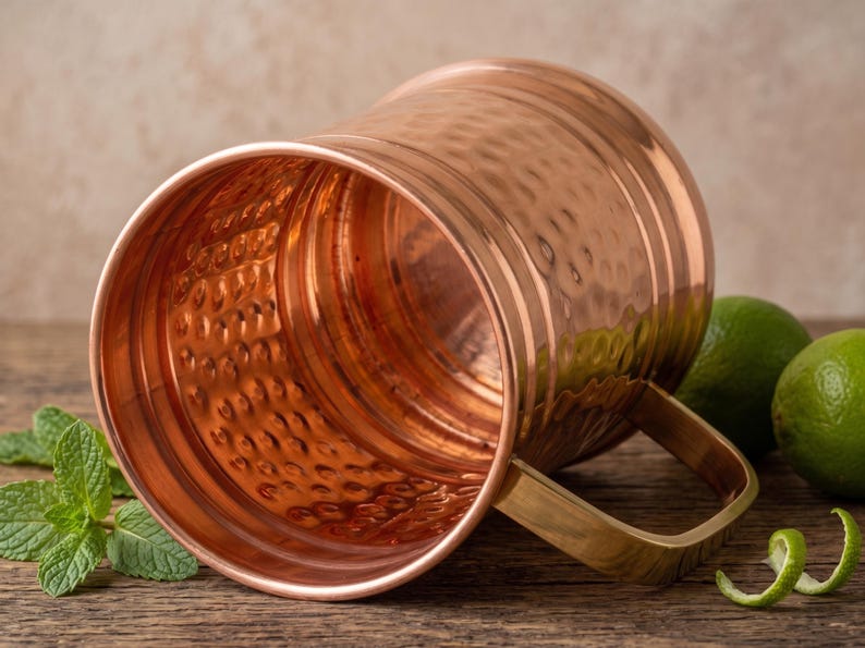 May include: A polished copper mug with a hammered texture on the exterior and interior surfaces. The mug features a smooth, flat-bottomed base and a sturdy, rectangular-shaped handle attached to the side, reflecting a warm metallic sheen.