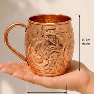 May include: A copper mug with an embossed floral pattern and a curved handle. The mug features a height of 10 cm and a width of 7 cm as indicated by the measurement markers. The exterior surface has a polished metallic finish with intricate detailing.