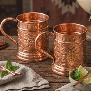 May include: Two polished copper mugs featuring embossed leaf patterns on the exterior, a smooth rim, and a sturdy handle. The mugs have a metallic finish and a cylindrical shape. A small bowl with lime wedges and mint leaves sits nearby.