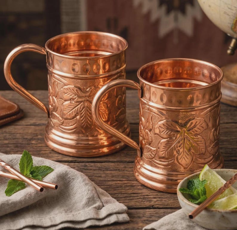 May include: Two polished copper mugs featuring embossed leaf patterns on the exterior, a smooth rim, and a sturdy handle. The mugs have a metallic finish and a cylindrical shape. A small bowl with lime wedges and mint leaves sits nearby.