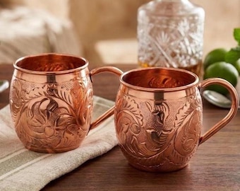 May include: Two round copper mugs featuring an intricate embossed floral pattern across the exterior surface. Each mug has a curved handle attached to the side. The metal has a polished, reflective finish that highlights the detailed engravings.