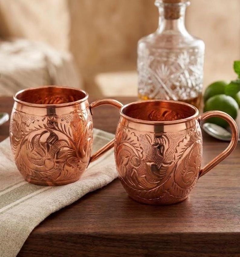 May include: Two round copper mugs featuring an intricate embossed floral pattern across the exterior surface. Each mug has a curved handle attached to the side. The metal has a polished, reflective finish that highlights the detailed engravings.