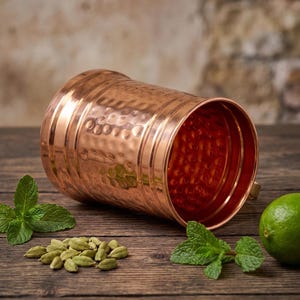 May include: A cylindrical copper mug with a hammered exterior texture and smooth horizontal bands. The mug is positioned horizontally, revealing its polished interior. Nearby are several green cardamom pods, fresh mint leaves, and a lime.