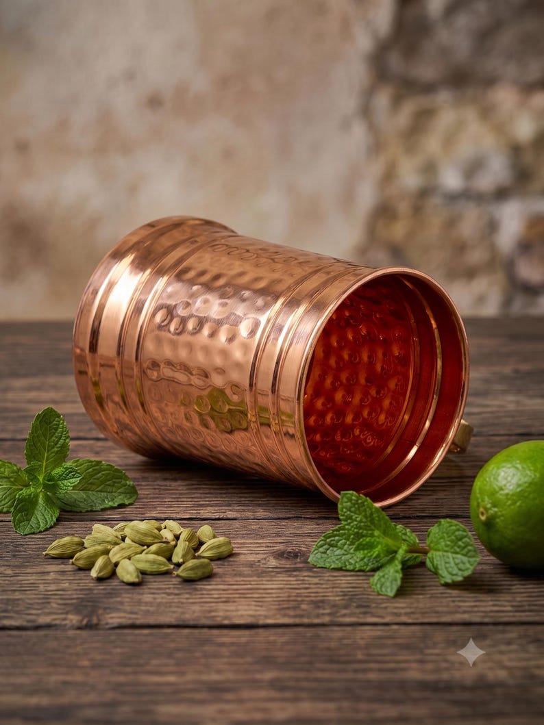 May include: A cylindrical copper mug with a hammered exterior texture and smooth horizontal bands. The mug is positioned horizontally, revealing its polished interior. Nearby are several green cardamom pods, fresh mint leaves, and a lime.