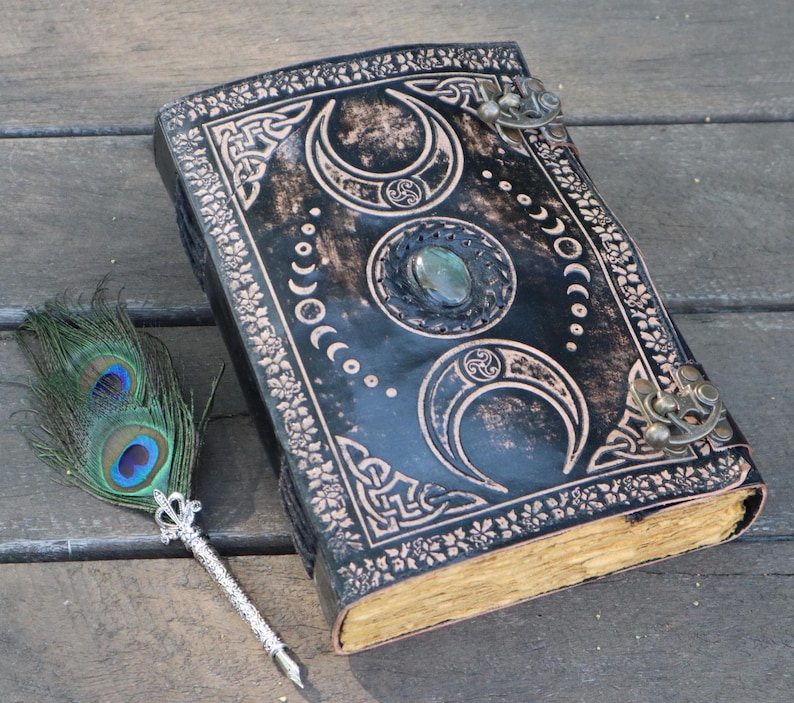May include: A black leather journal with a triple moon design, a gemstone embellishment, and two brass clasps. A peacock feather pen is lying next to the journal.