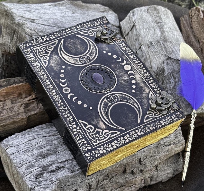 May include: Black leather journal with a gold and silver moon and Celtic knot design. The journal has a purple gemstone and metal clasps.