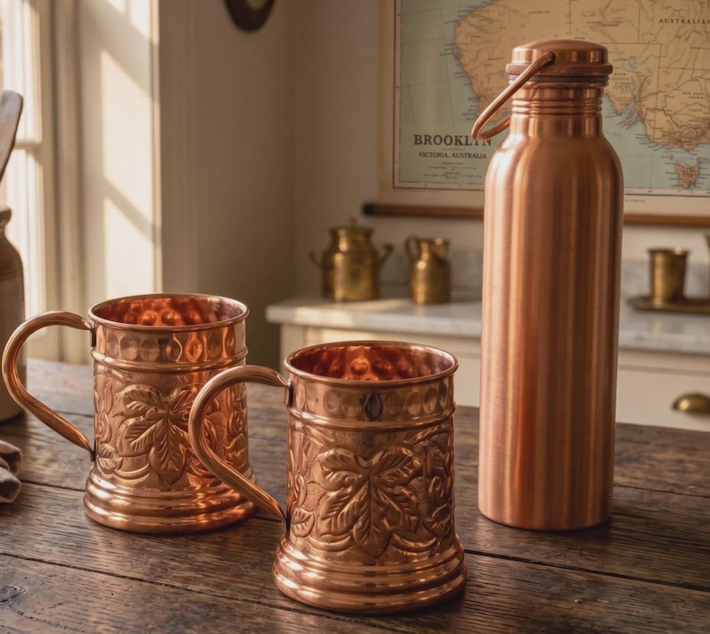 May include: A set of two copper mugs with embossed leaf patterns and a matching tall copper water bottle with a handle on the lid. The mugs feature a cylindrical shape with a sturdy side handle, while the bottle has a smooth, polished finish.
