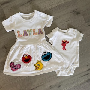 May include: A white short-sleeved dress featuring the name LAYLA in glittery letters and patches of Elmo, Cookie Monster, Abby Cadabby, and Big Bird on the skirt, alongside a white short-sleeved bodysuit with an embroidered red Elmo patch.